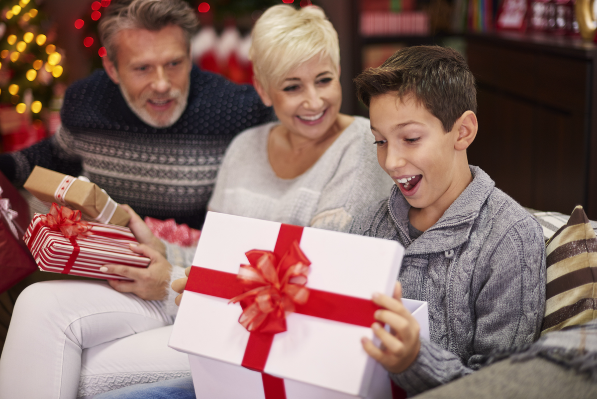 A young boy looking thrilled and surprised as he opens a large white gift box with a bright red bow, surrounded by his smiling parents.