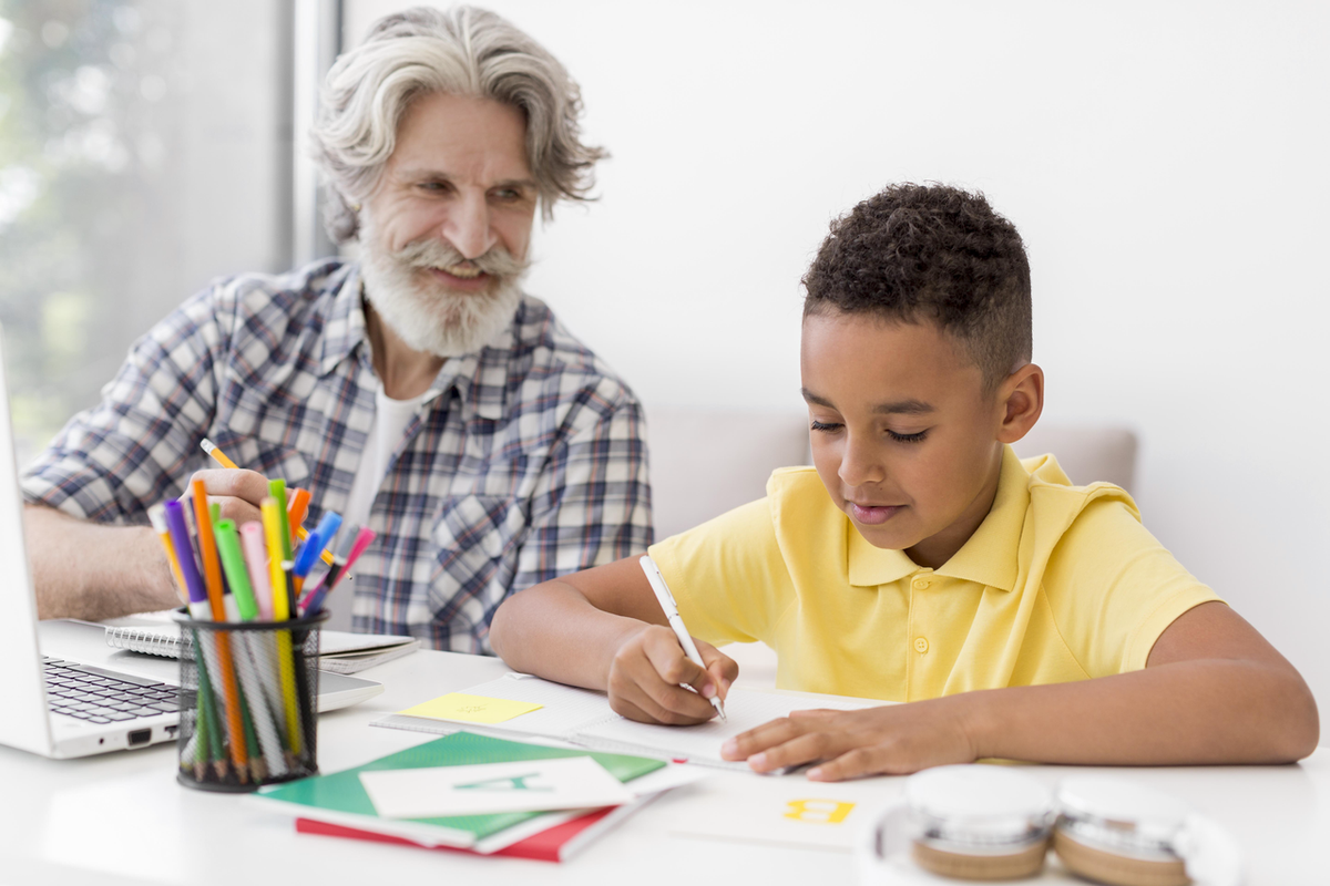 A supportive grandfather watches as a student in a yellow shirt writes in his book. This moment of guidance reflects the enduring love that a grandfather shows when gifting a symbolic botanical bracelet.