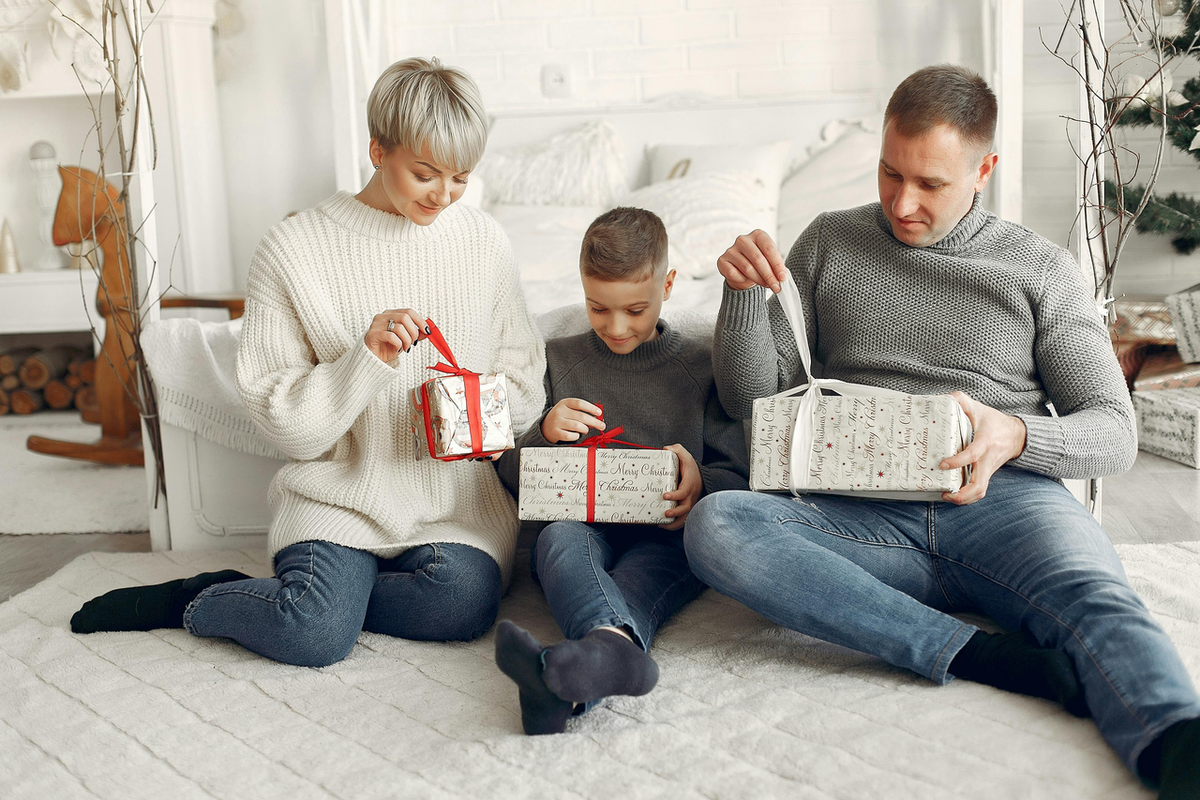 A family sitting on a white rug in a cozy, decorated room, while smiling and opening wrapped family gifts.
