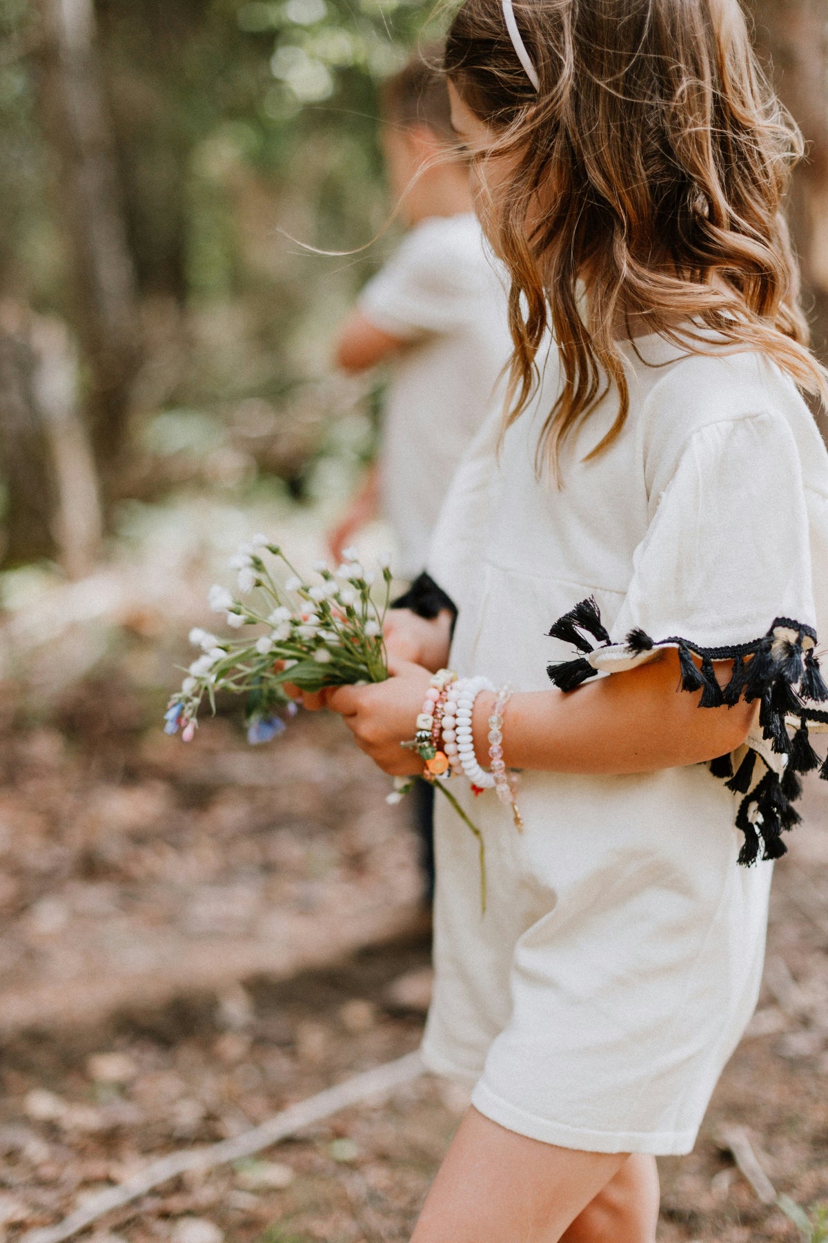 A girl in a light dress stands in a wooded area holding wildflowers, wearing colorful flower jewelry bracelets. The peaceful outdoor scene reflects SARAH'S WHISPER’s nature-inspired designs.